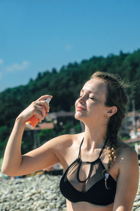 Μια όμορφη νεαρή γυναίκα με μαγιό αναζωογονεί και ενυδατώνει το πρόσωπό της με facial mist ενώ βρίσκεται στην παραλία κάτω από τις λαμπερές ακτίνες του καλοκαιρινού ήλιου