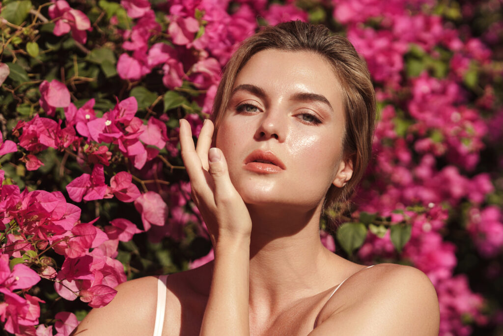 Όμορφη ξανθιά γυναίκα με λείο ενυδατωμένο δέρμα και φυσικό μακιγιάζ, καθώς ποζάρει στον ήλιο με φόντο έναν θάμνο με ροζ λουλούδια.