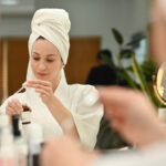 γυναίκα με μπουρνούζι εφαρμόζει σταγόνα αιθέριου ελαίου στο πίσω μέρος του χεριού της, κάνοντας την καθημερινή της ρουτίνα στο σπίτι.